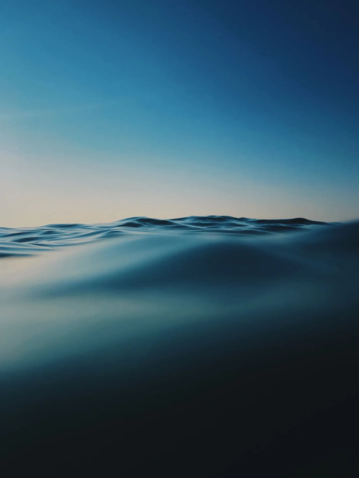 A low-angle, water-level close-up shot of deep blue ocean swells under a clear, transitioning sky, evoking a sense of vastness and quiet isolation