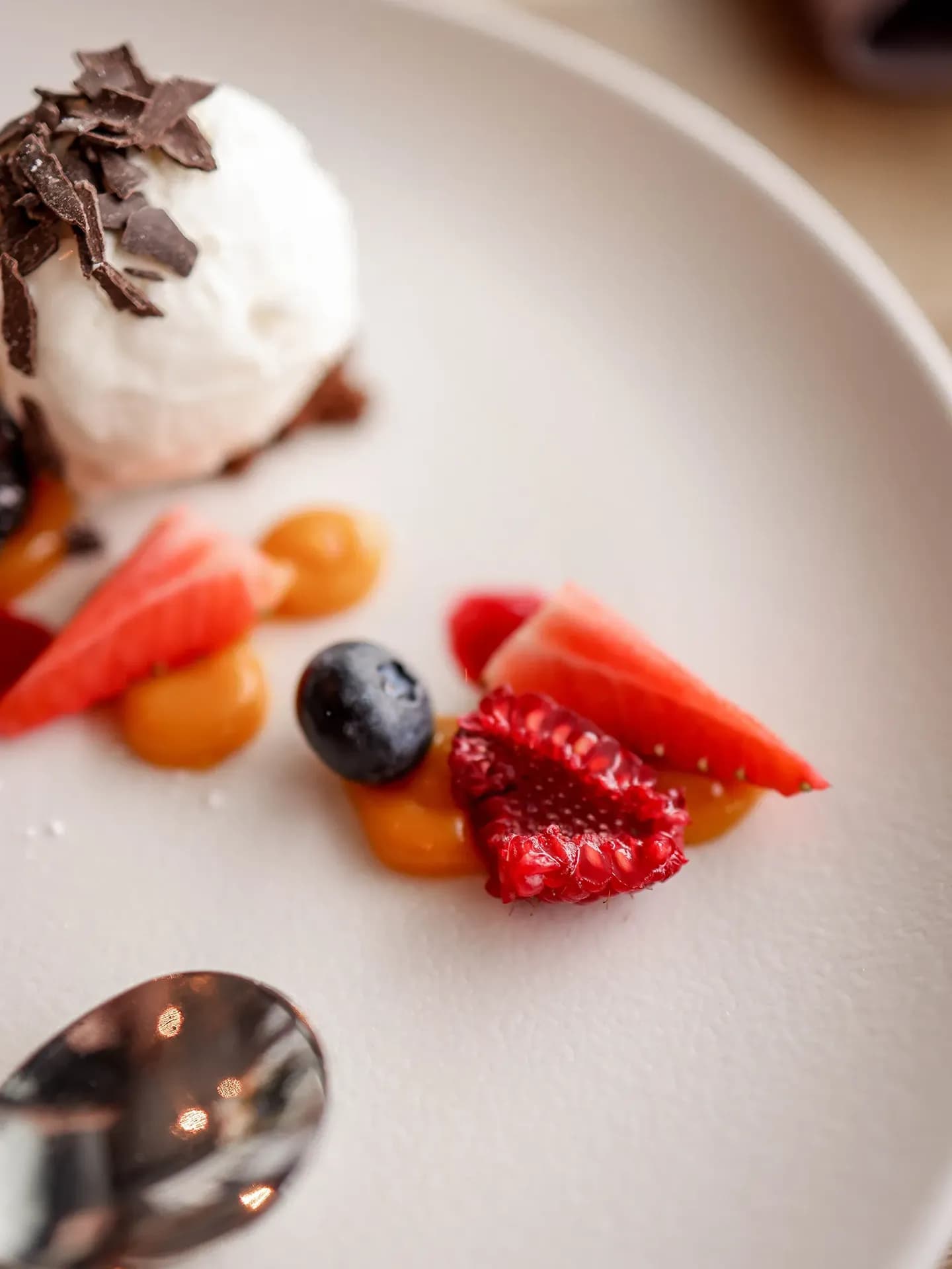 A close-up, high-angle shot of an elegant dessert on a matte ceramic plate, featuring a scoop of vanilla ice cream topped with chocolate shavings, paired with fresh raspberries, blueberries, strawberries, and artistic dollops of golden coulis