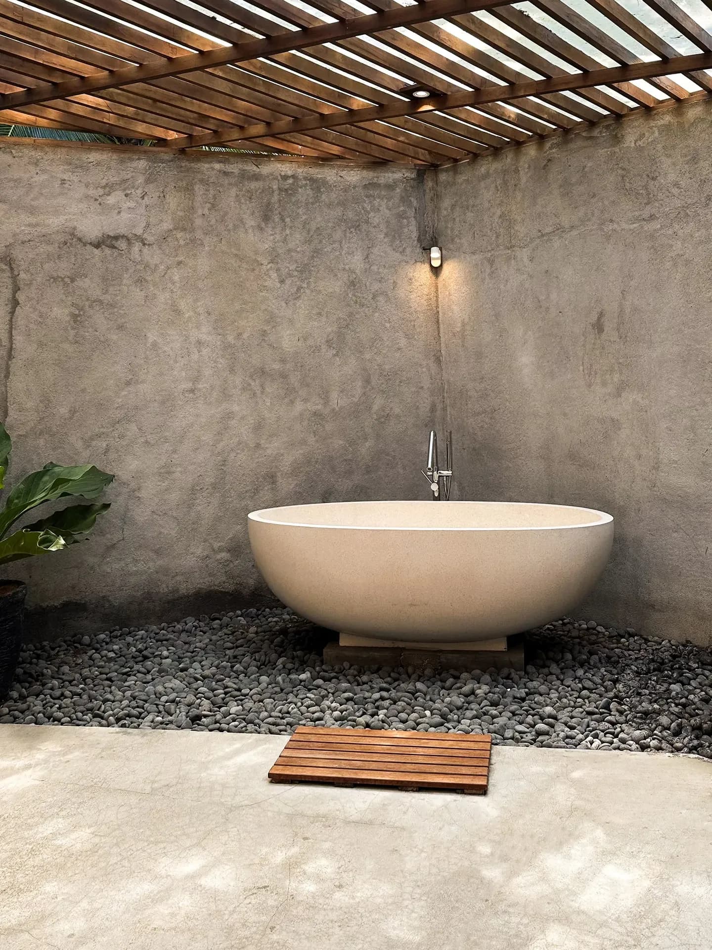 A sophisticated outdoor bathroom sanctuary showcasing a round, cream-colored stone soaking tub set over grey river pebbles, framed by raw concrete walls and a structural wooden slat pergola roof